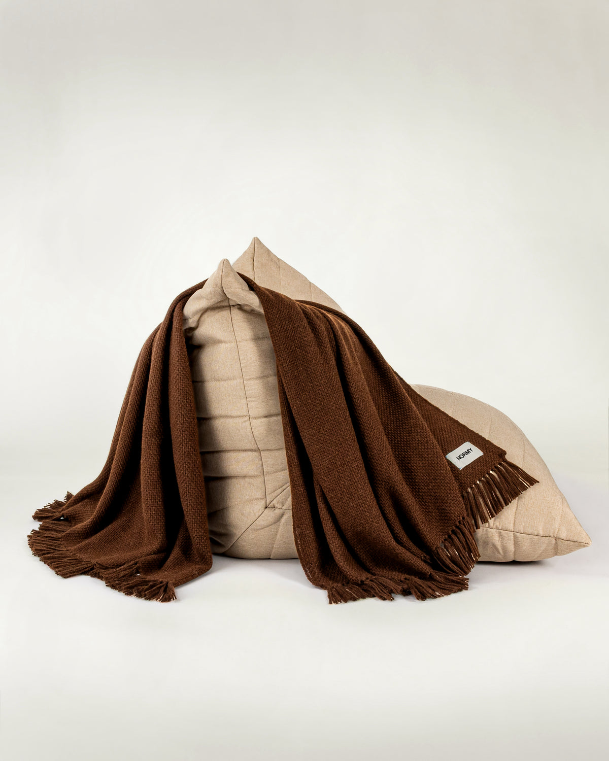Normy Alpaca Blanket in cocoa draped over a beige quilted cushion, set against a plain light gray studio background.