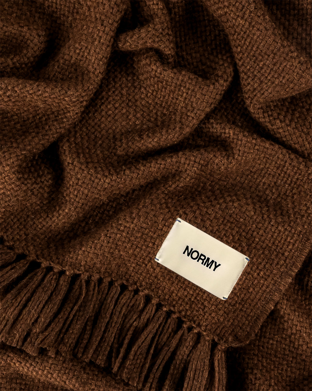 A close-up of Normy Alpaca Blanket in cocoa and a small cream fabric label reading NORMY.