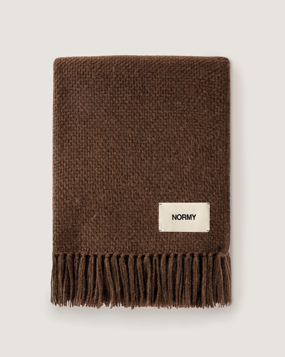 Normy Alpaca Blanket in cocoa with a cream-colored rectangular label that reads 'NORMY', shown flat against a light gray background.