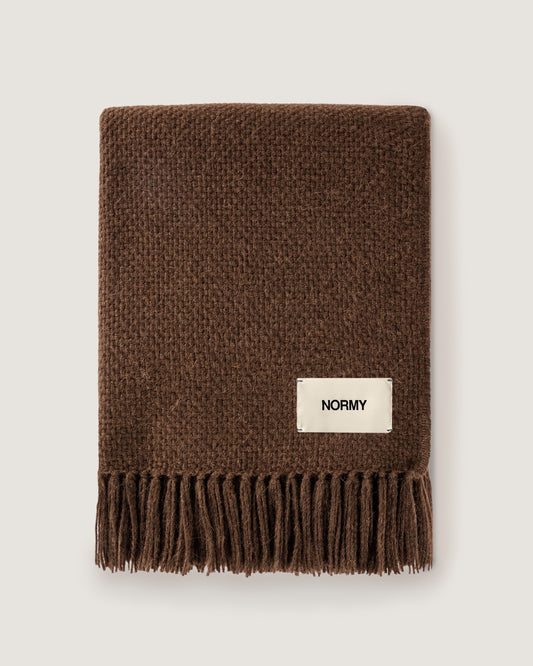 Normy Alpaca Blanket in cocoa with a cream-colored rectangular label that reads 'NORMY', shown flat against a light gray background.