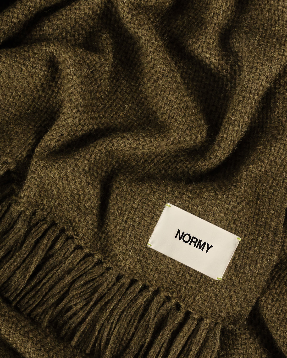 Close-up of Normy Alpaca Blanket in olive with a white fabric label reading 'NORMY' and fringe along one edge.