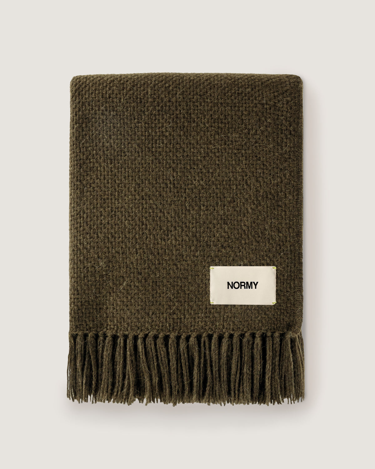 Normy Alpaca Blanket in olive with a cream-colored 'NORMY' brand label, displayed flat against a light gray background.