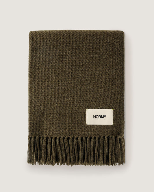 Normy Alpaca Blanket in olive with a cream-colored 'NORMY' brand label, displayed flat against a light gray background.